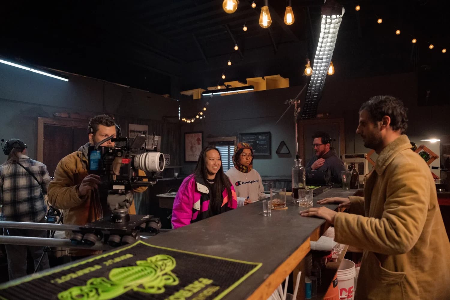 Olivia Wong on set behind a bar, watching a take, with crew and lighting rig around her