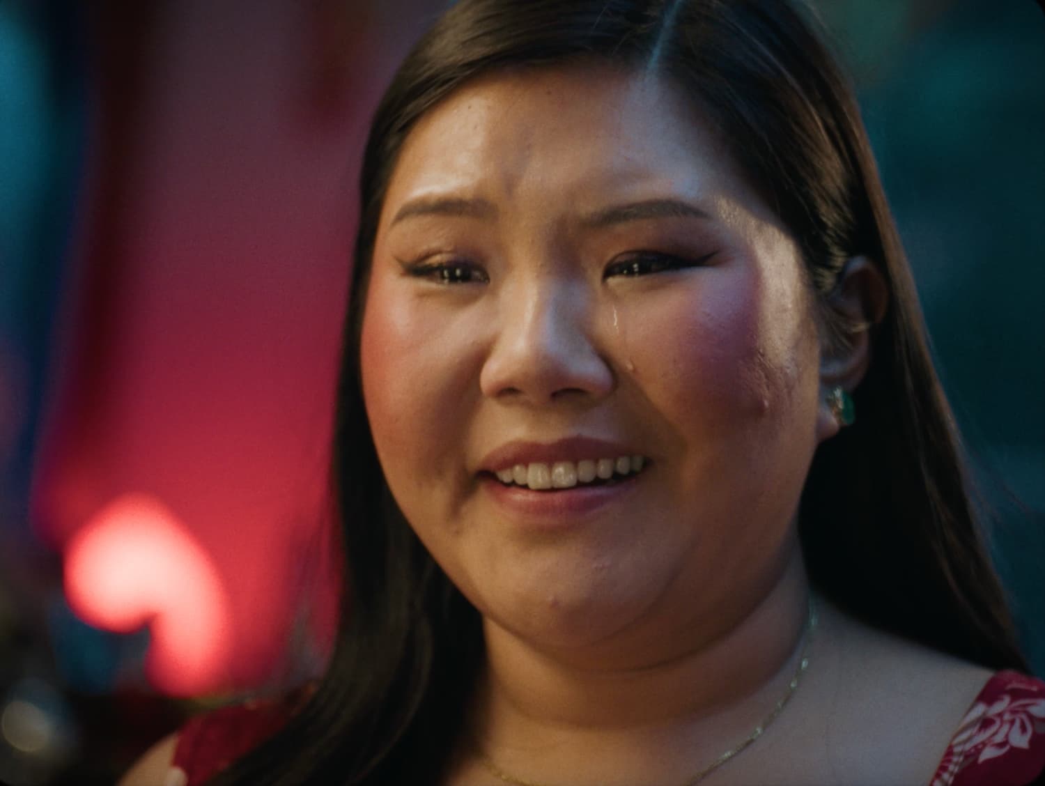 A young woman smiling through tears in close-up, a red heart-shaped light glowing out of focus behind her — a still from the Kohl's spec commercial