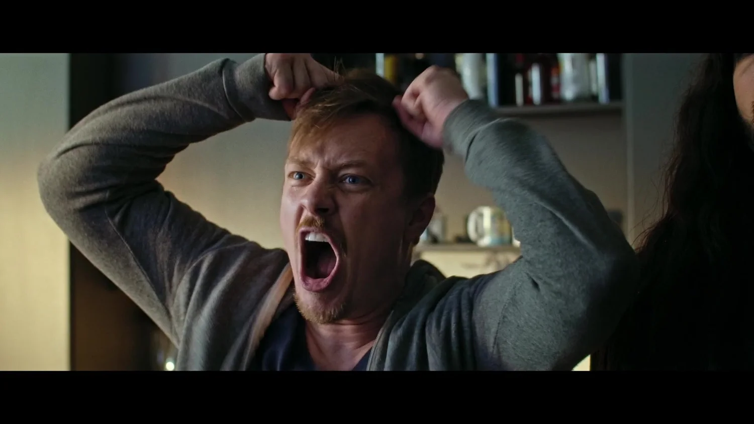 A man screaming with both fists gripping his hair in a kitchen, another figure just visible at the edge of frame — a still from Madness Incarnate