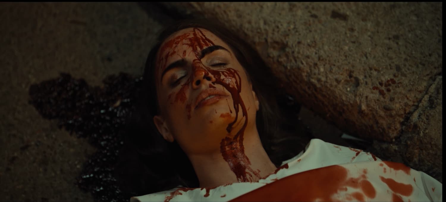 A woman lying on concrete with her eyes closed, blood streaking her face and white shirt and a dark pool spreading beside her head — a still from the horror short You're IN